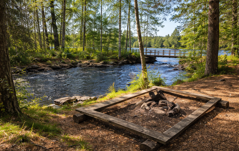 Lugn skogsmiljö vid en flod med en eldstad och en bro i bakgrunden.