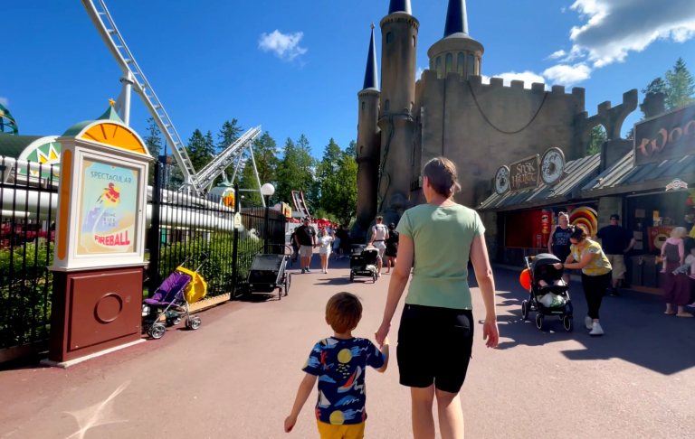 En vuxen och ett barn går mot ett sagoslott i en nöjespark. Många besökare syns i bakgrunden.