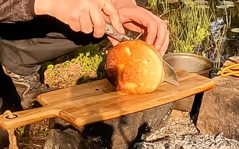 En hand som skär bröd på en träskärbräda vid en lägereld.