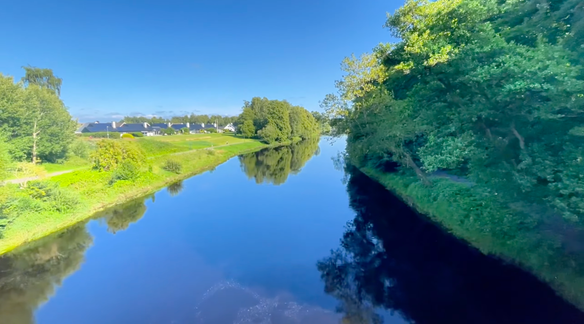 Ätran vid Vallarnas Friluftsområde Spegelblanka vattenytor omges av grönska och träd under en klarblå himmel.