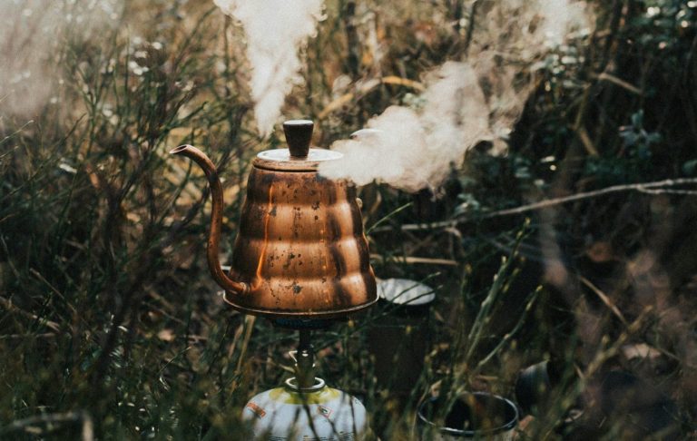 En gammaldags vattenkokare med rök som stiger, omgiven av grön vegetation.