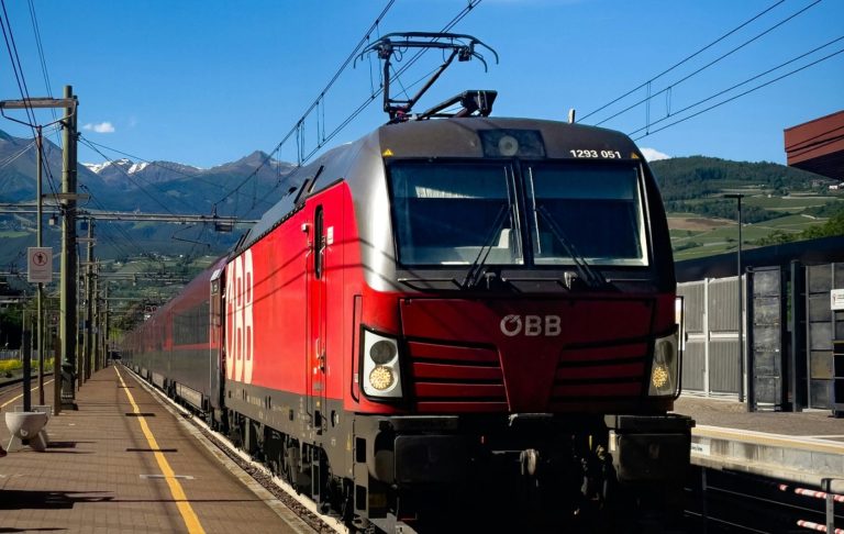 Röd elektrisk tåg bakom plattform med berg i bakgrunden.