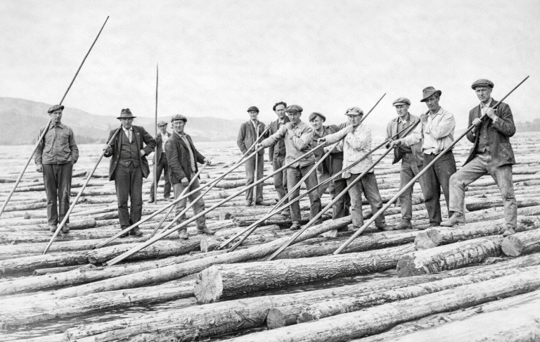 Grupp män som arbetar med stockar, hållande pinnar, vid en flod eller insjö.