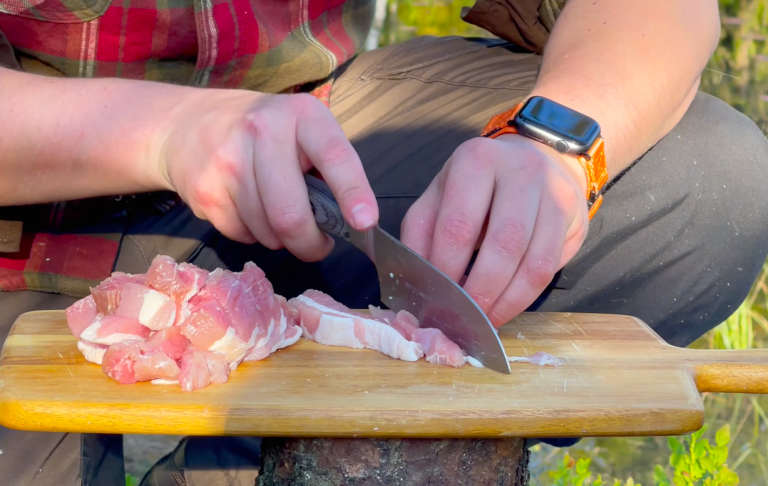 En person skär kött på en skärbräda med en kniv i en utomhusmiljö.