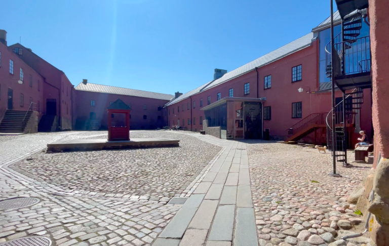 Gård med stenlagd gård och röd byggnad under klarblå himmel.