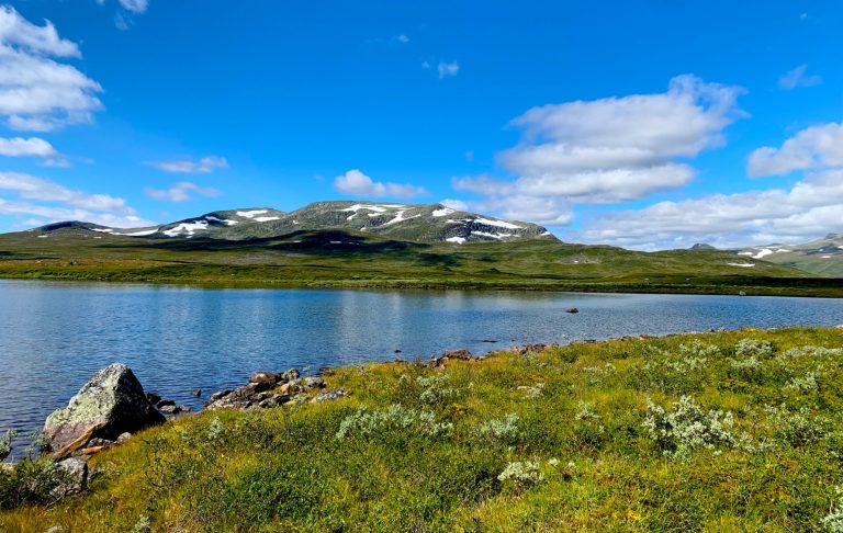 Fjällandskap med en spegelblanka sjö och fjäll täckta av snö under en blå himmel.