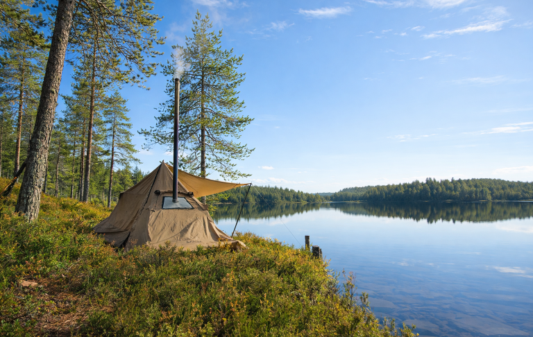 Hot tent med absid Kamin Tält vid en spegelblanka sjö med skogiga omgivningar och klarblå himmel.