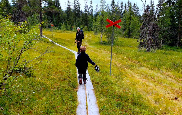 Två personer promenerar på en stig i en skog med röd varningsskylt.