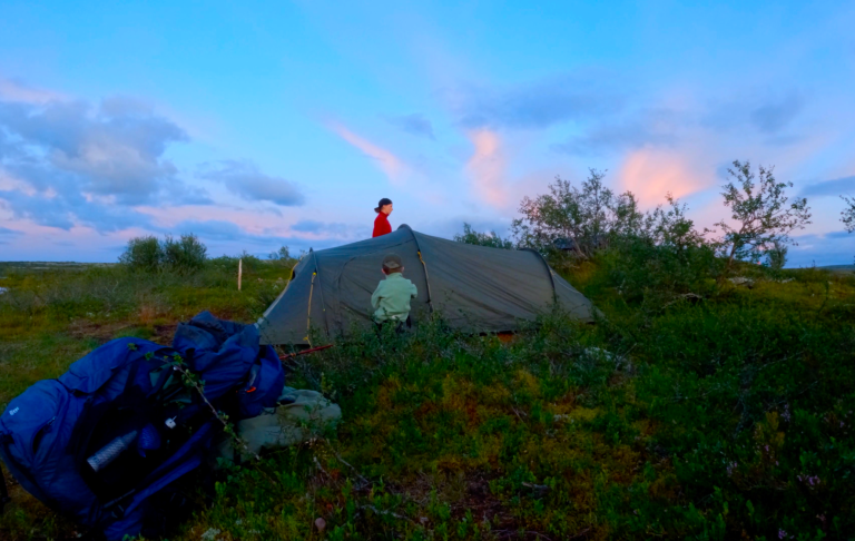 Tält på en grön äng med två personer vid solnedgången.