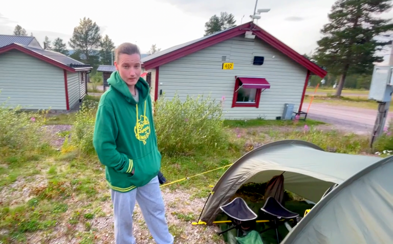 Ung tjej står vid ett tält nära en rödmålad stuga i naturen.