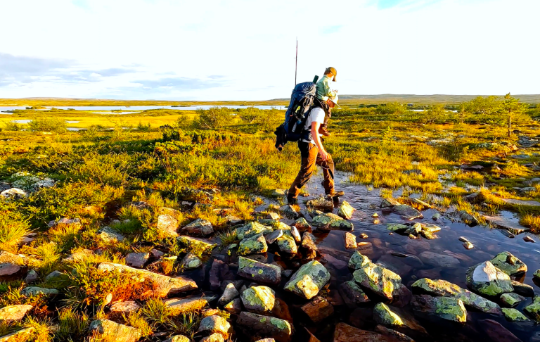 En person vandrar med ryggsäck över stenar i ett öppet, grönskande landskap.