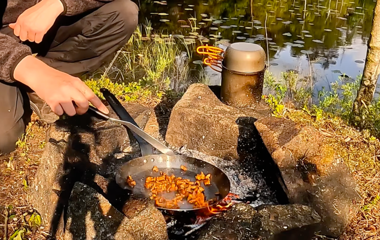 En person lagar mat i en stekpanna över öppen eld vid en sjö.