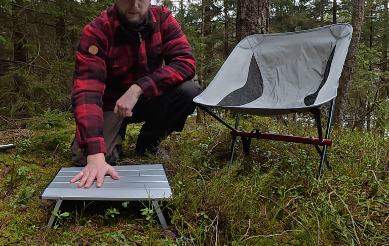En person i röd-rutig jacka placerar en bärbar bänk nära en campingstol i skogen.
