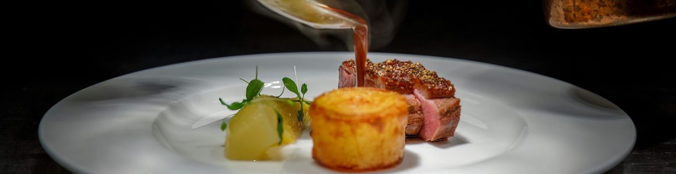 Servering av en tallrik med kött, potatisgratäng och grönsaksgarnityr.