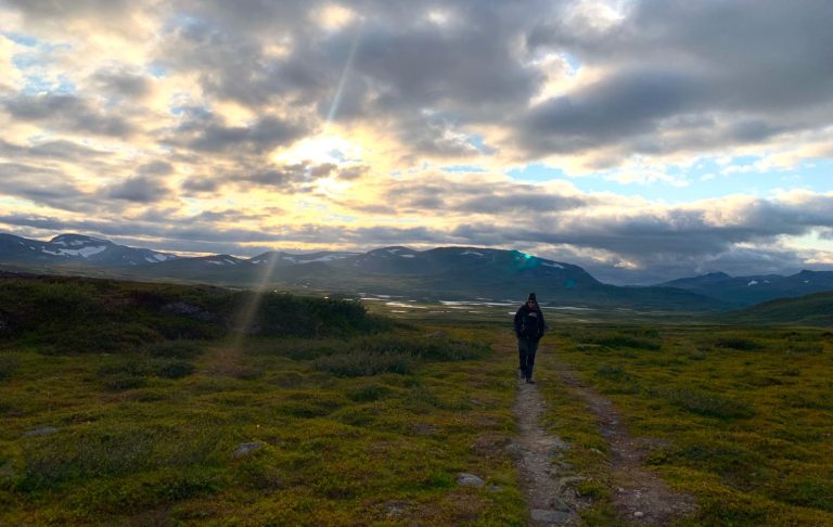 Person med barn på magen som går längs en stig i en grönskande landskap under molnig himmel.