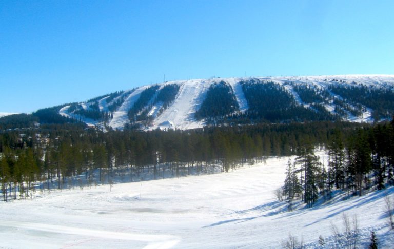 Snöklädd bergskedja med granar och en frusen sjö i förgrunden. Klara blå himmel.