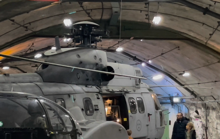 HKP 10 Super Puma på Aeroseum En militärhelikopter står inuti en hangar med belysning och människor i bakgrunden.