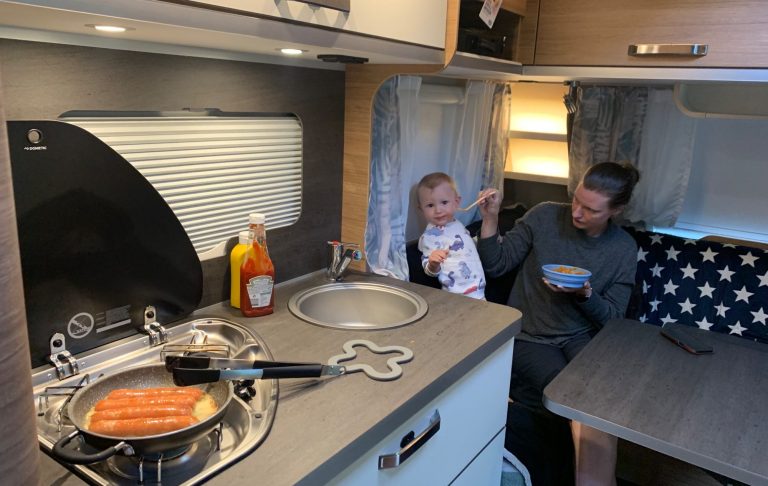 En förälder lagar mat i en husvagn medan ett barn leker vid bordet.
