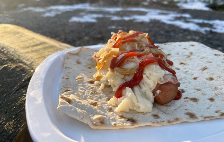 Korv med potatismos, lök och ketchup på ett tunnbröd. Solnedgång och en grusstig med snö i bakgrunden.