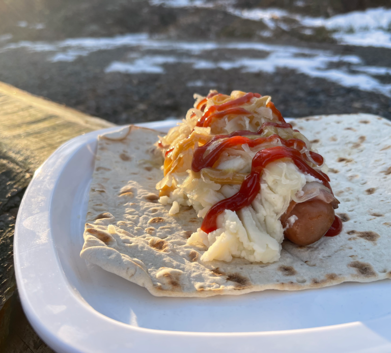 Korv med mos, lök och ketchup i ett tunnbröd på en tallrik. Snöigt landskap i bakgrunden.