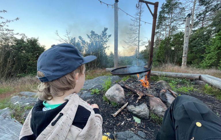 Barn som sitter vid en lägereld, omgiven av natur och rök i luften.