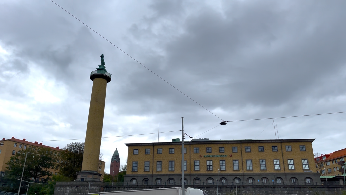 Sjöfartsmuseet Kägelbrunnens pelarstaty med grå himmel och byggnad i bakgrunden.