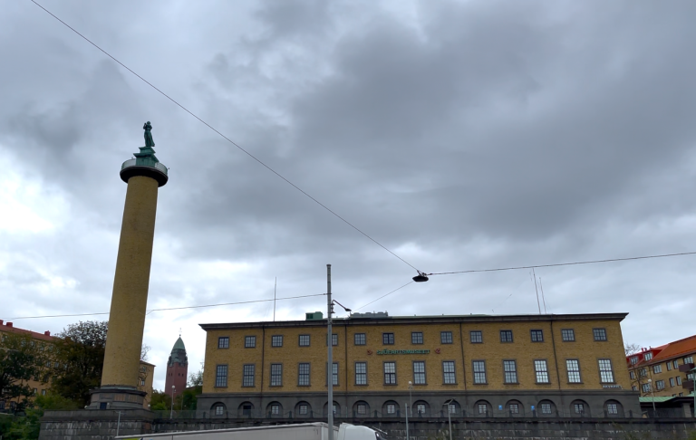 Ett gult byggnad med en hög pelare och en staty under en molnig himmel.