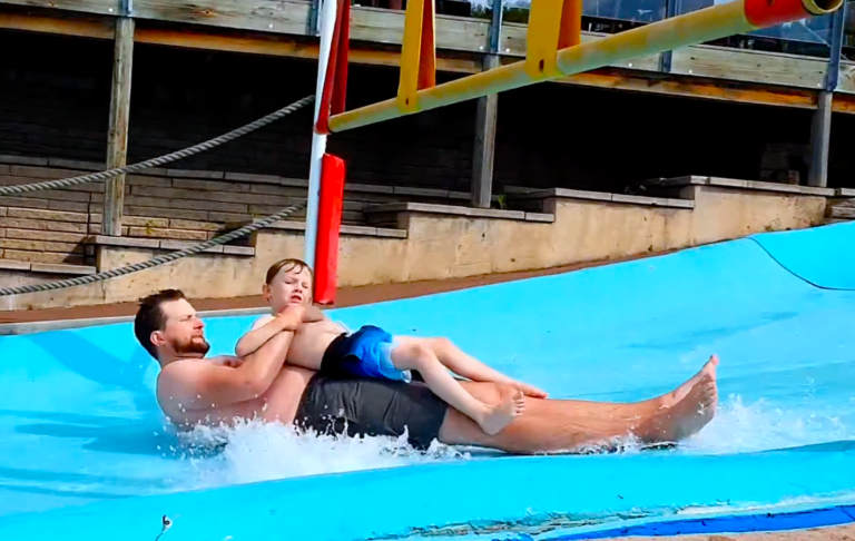 Man and boy sliding down a water slide, splashing in the water below.
