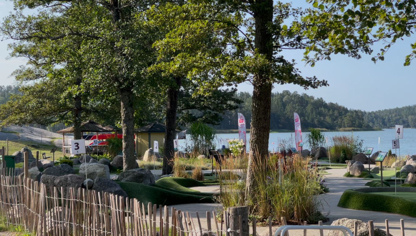 Minigolfen Grönskande park vid en sjö med träd, bänkar och flaggor i bakgrunden.